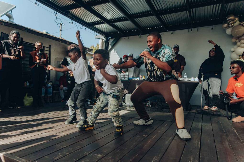 two young boys dance on a deck beneath a pergola while a 29-year-old rapper eggs them on