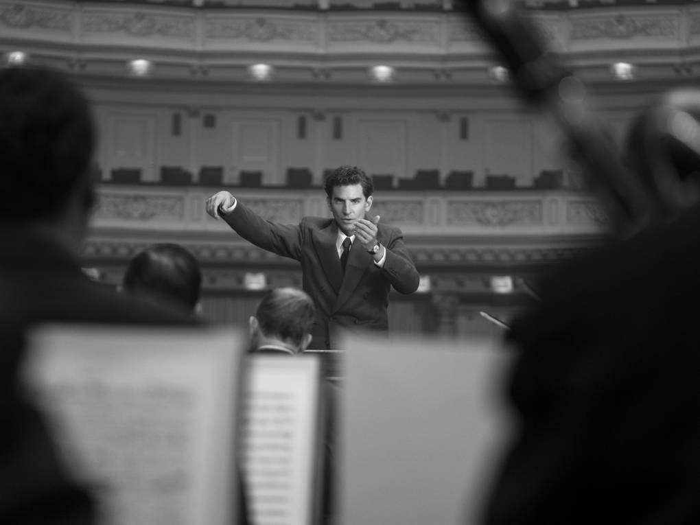 black and white image of a conductor seen from several rows back in orchestra, over music stands