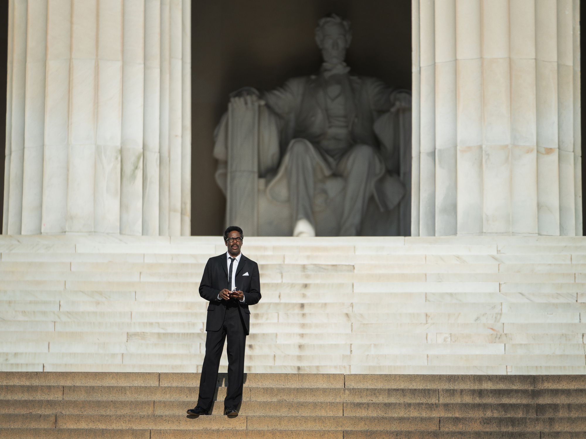 A Black man in a black suit stands on the Lincoln Memorial steps, small against the architecture and memorial