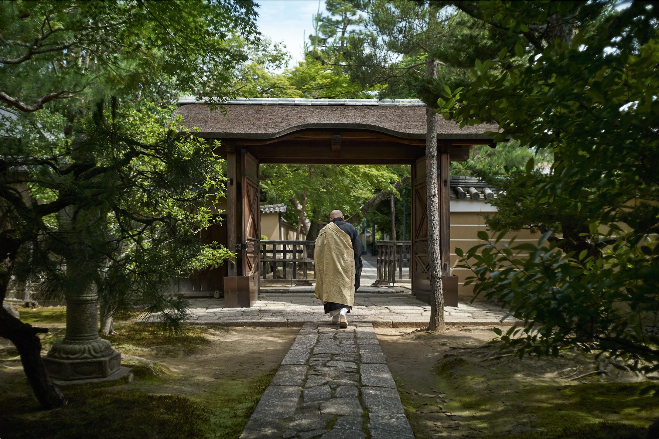 Man in robe walks down stone path under gateway, lush trees surrounding