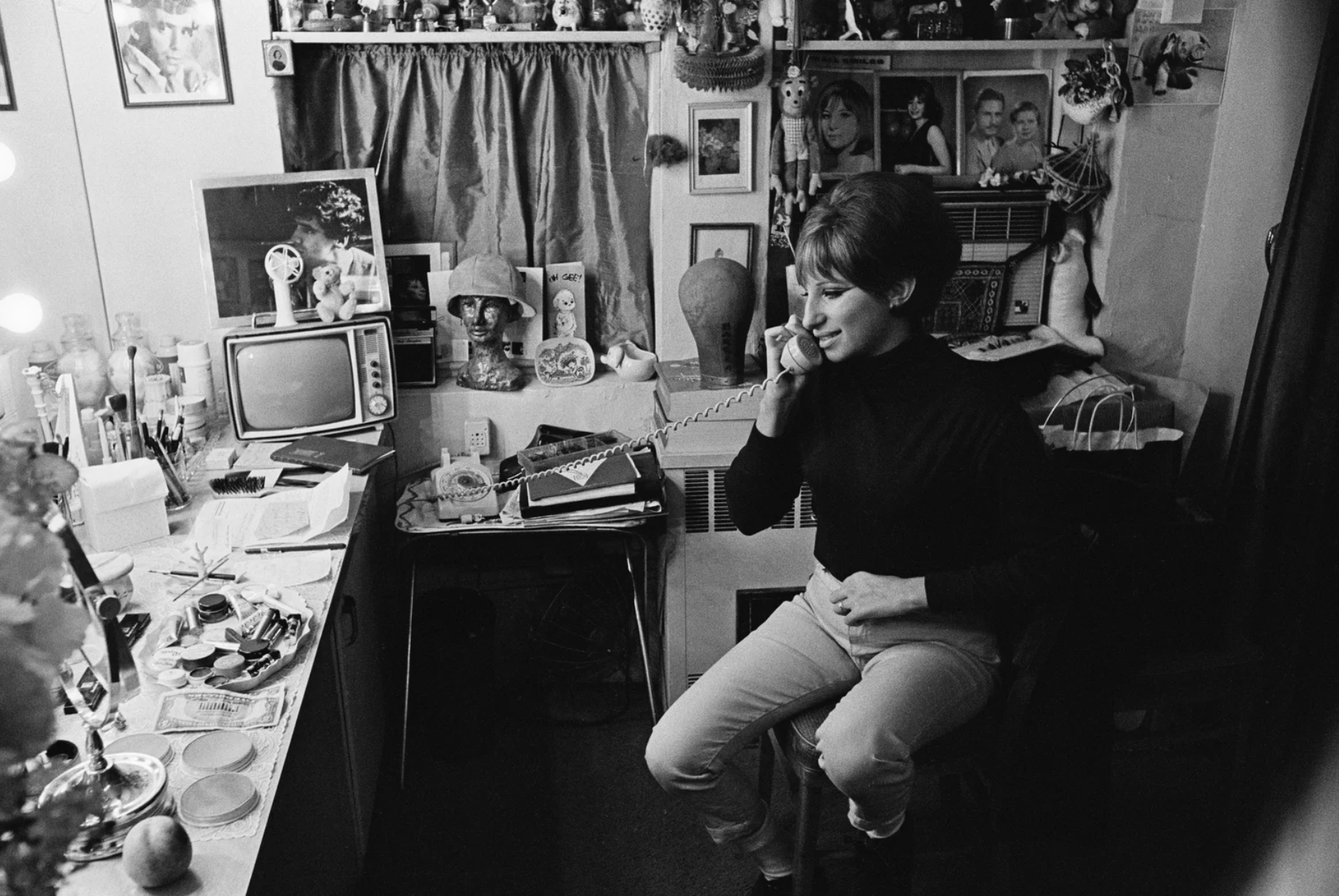 A young woman, casually dressed, talks on a landline telephone in a dressing room, a table scattered with make-up in front of her.