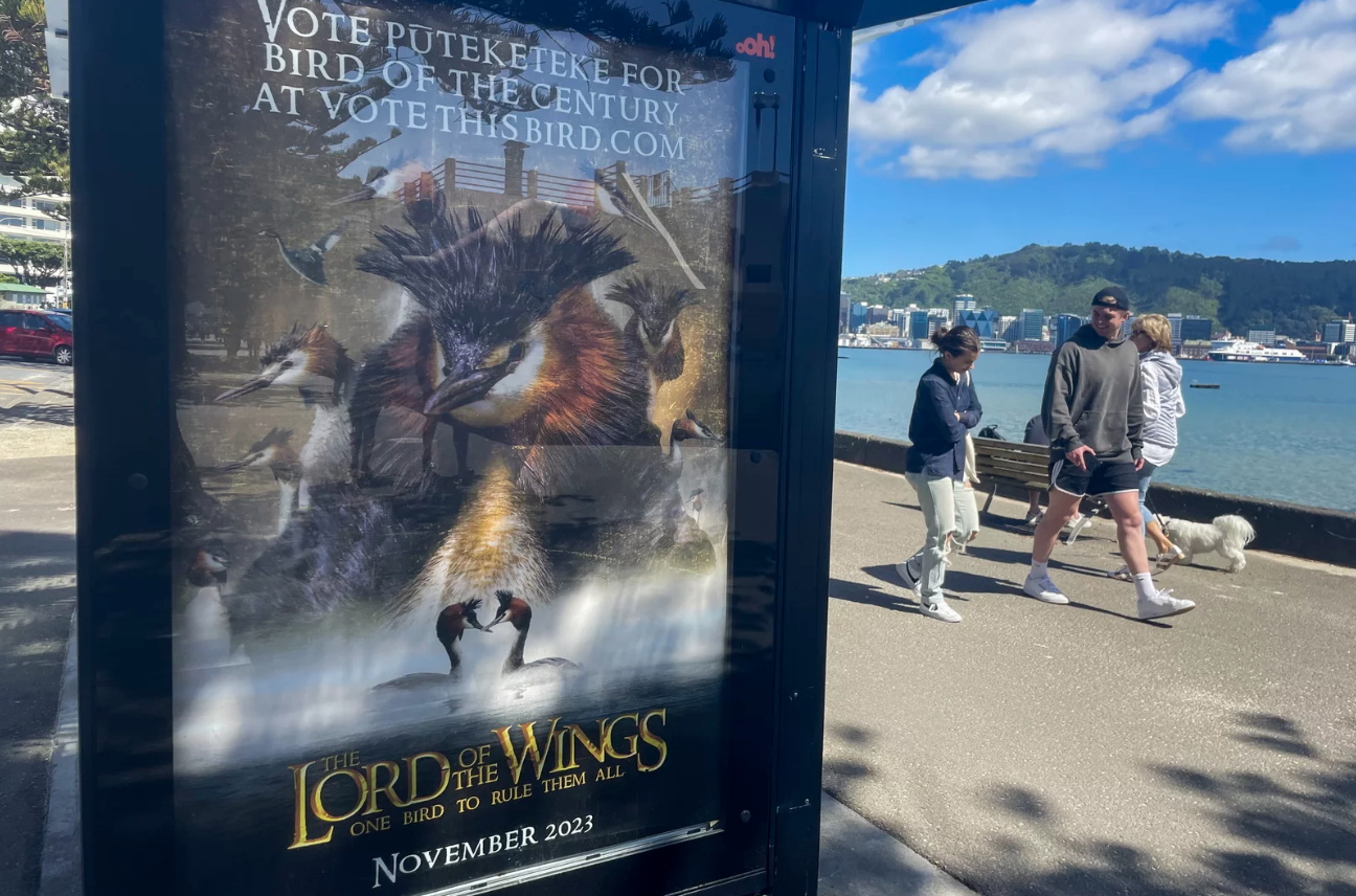 A couple walks past a bus billboard depicting a parody of Lord of the Rings featuring unusual birds. "Lord of the wings," it reads. "One bird to rule them all."