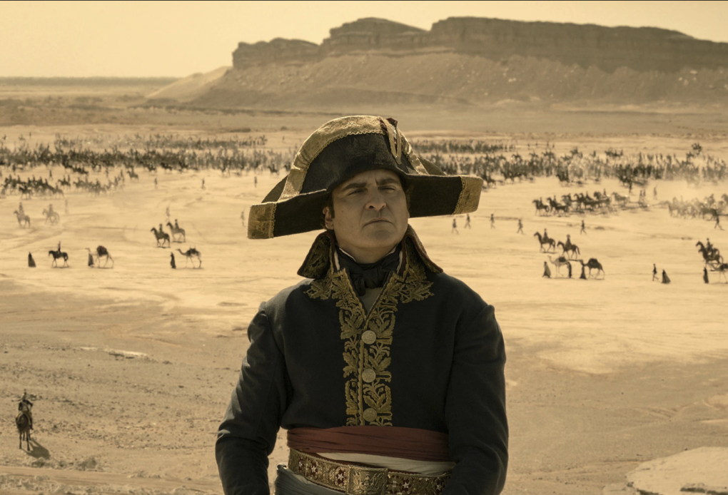 A white man in 18th century military uniform and hat stands on high ground. Behind him, there is a desert landscape covered in thousands of troops and horses.