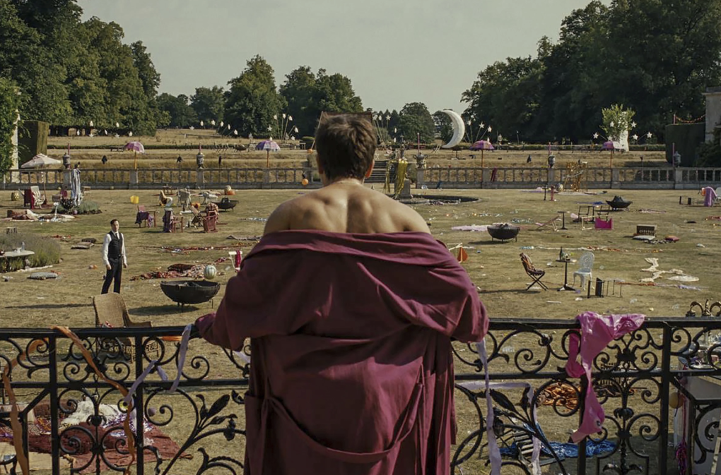A man with muscular shoulder exposed from a robe stands at a grand railing and overlooks a huge garden. It is strewn with detritus and trash from some kind of wild party.