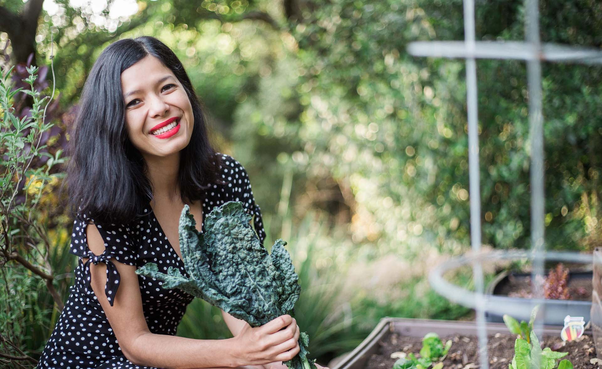 A woman seated outdoors next to a garden bed holds up a stalk of kale.