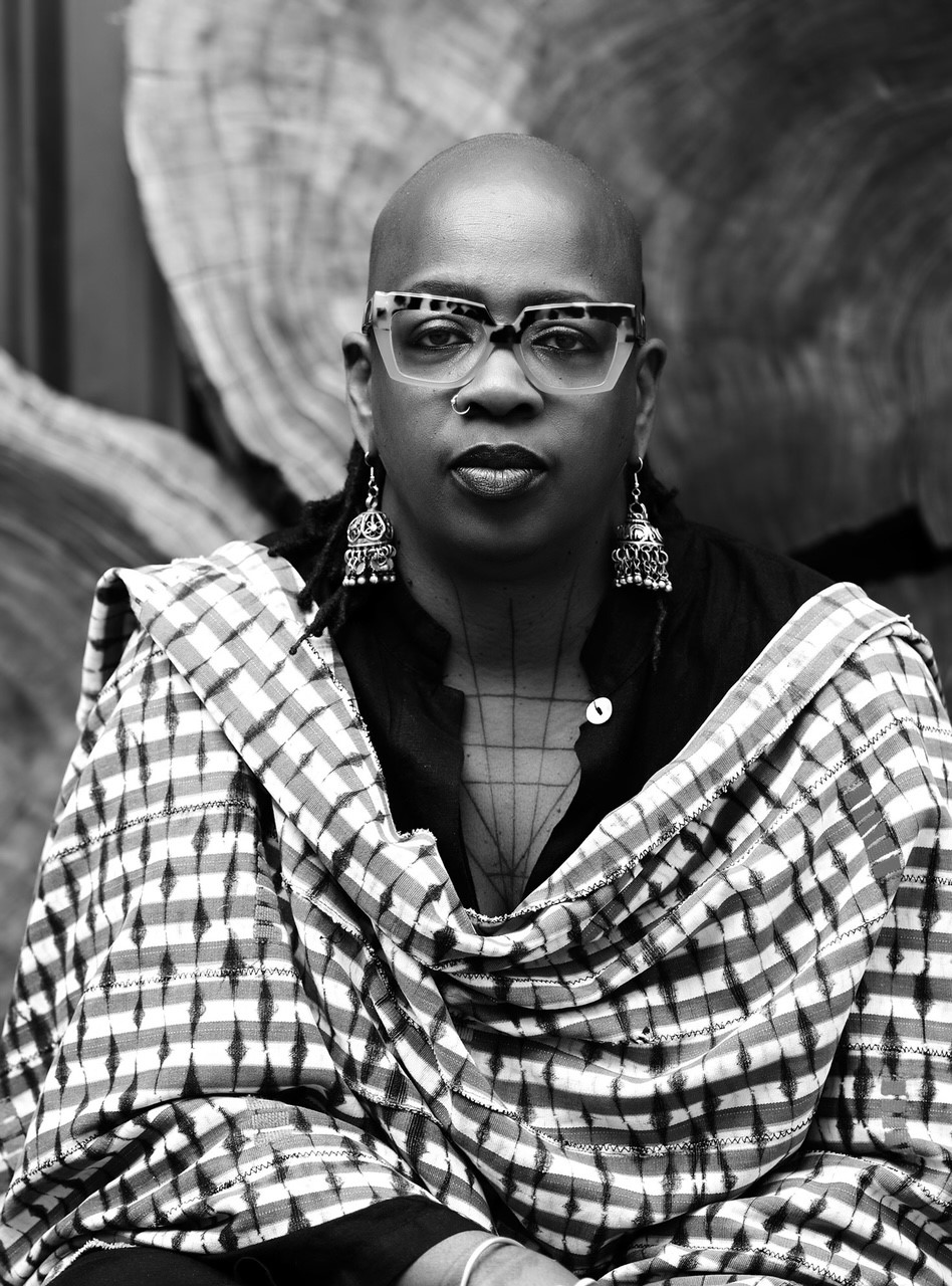 A black-and-white portrait of a female artist with a shaved head and glasses draped in a shawl.