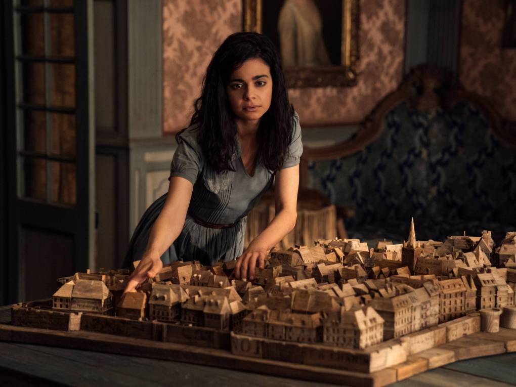 A young woman with long brown hair leans over the scale model of a town, touching it. She is wearing a blue dress and standing in a room with old-fashioned wallpaper and a grand fireplace.