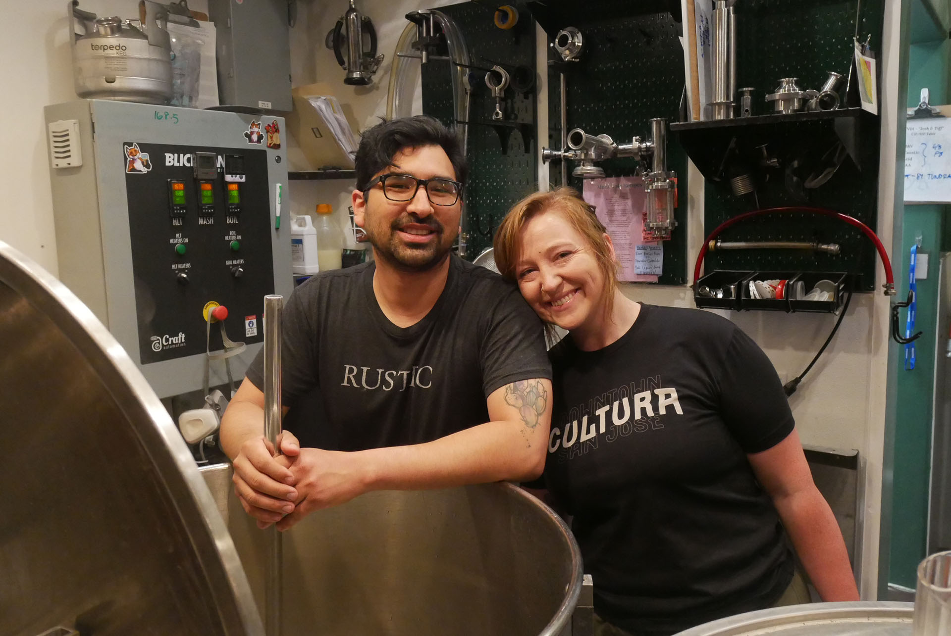 A man and woman in black T-shirts pose for a photo in front of a large kettle for brewing beer.