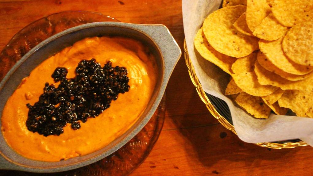A bowl of bright-orange vegan cashew "queso" topped with salsa macha, with a basket of tortilla chips on the side.