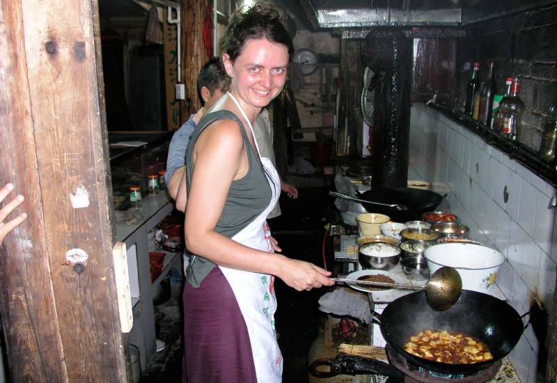 A young white woman in an apron cooks a dish in a wok in a traditional Chinese kitchen.