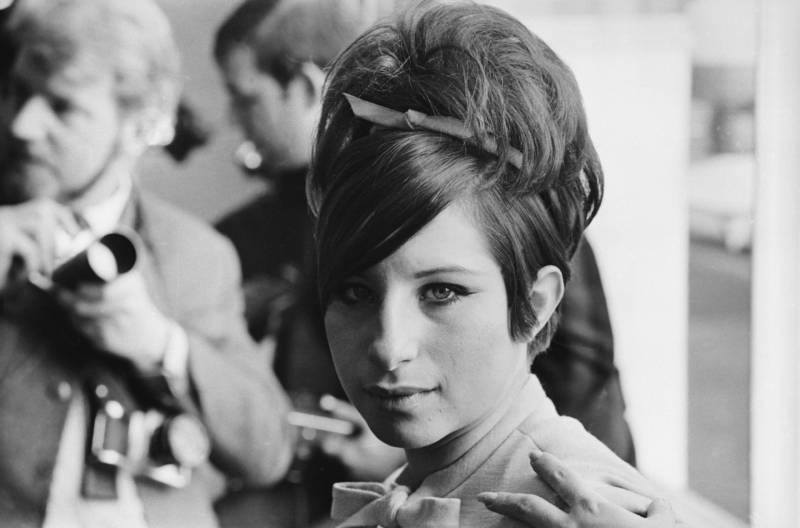 A young woman wearing a high necked blouse looks pensive. Her hair is in a small beehive and she's wearing distinctive black eyeliner.