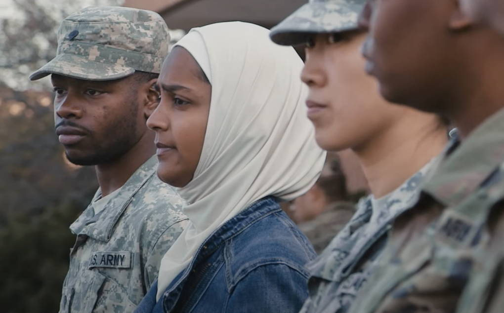 A woman of color wearing a white hijab and blue denim jacket lines up next to men and women wearing military uniforms.