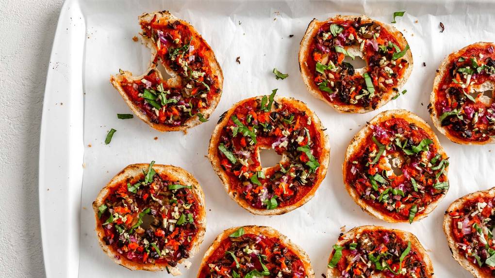 A tray of miniature pizza bagels topped colorfully with chopped herbs and vegetables.