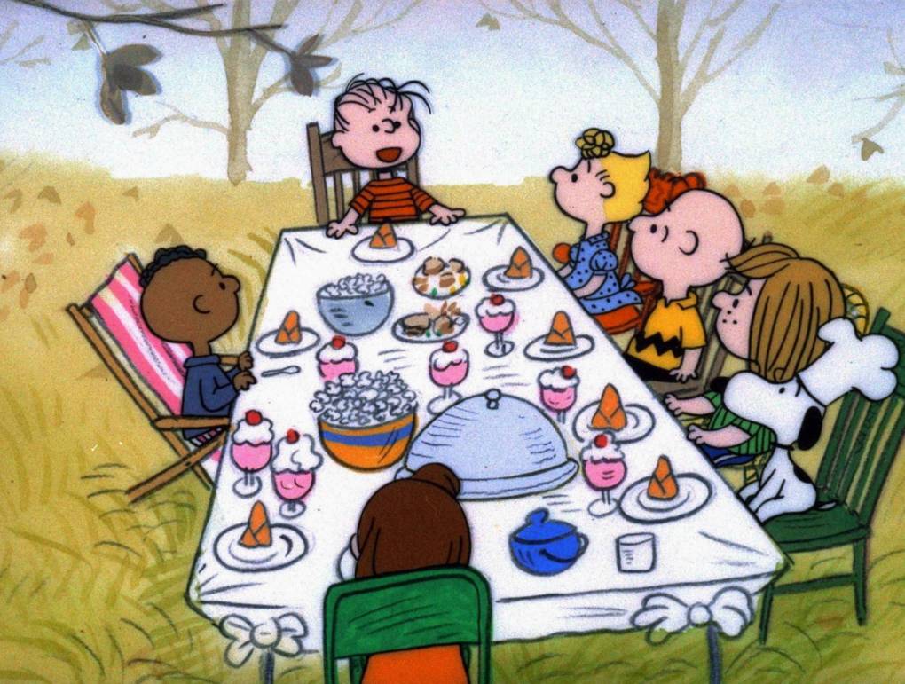 A scene from animated film 'A Charlie Brown Thanksgiving' featuring a group of white children sitting at the ends and on one side of a dinner table. A Black child is sitting alone on the opposite side of the table.
