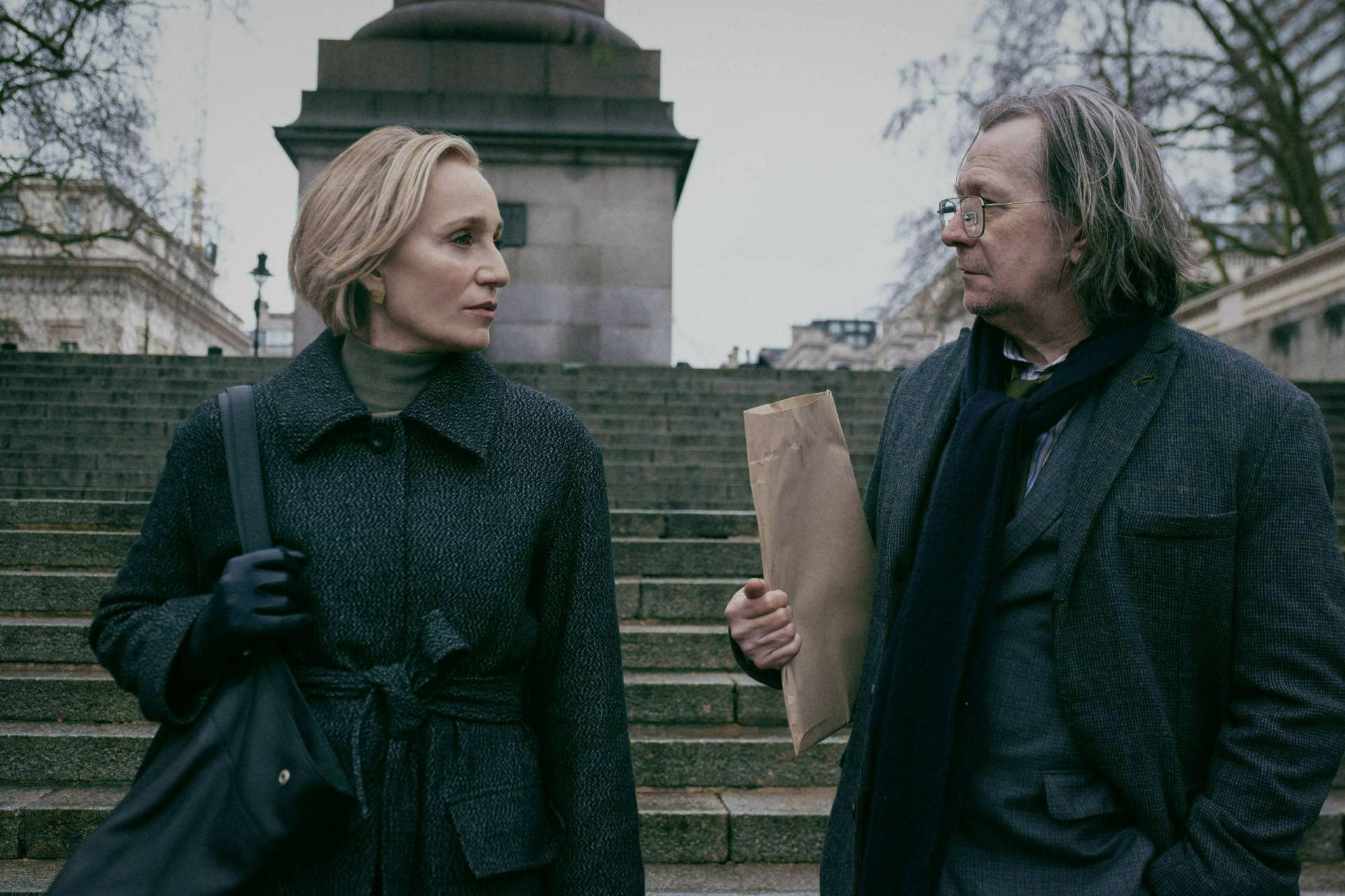 A slender white woman with a blonde bob stands at the bottom of a set of stone stairs. She is looking at an unkempt white man with long hair who is holding a brown envelope. The park they are standing in looks cold.