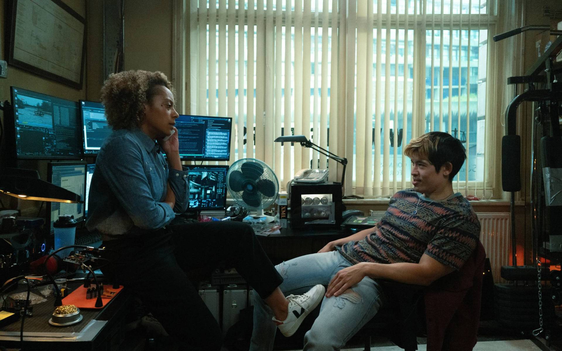 A young man wearing jeans and a T-shirt sits slouched in an office chair facing a young Black woman who is perched on a desk. Between them is computer equipment and a fan.