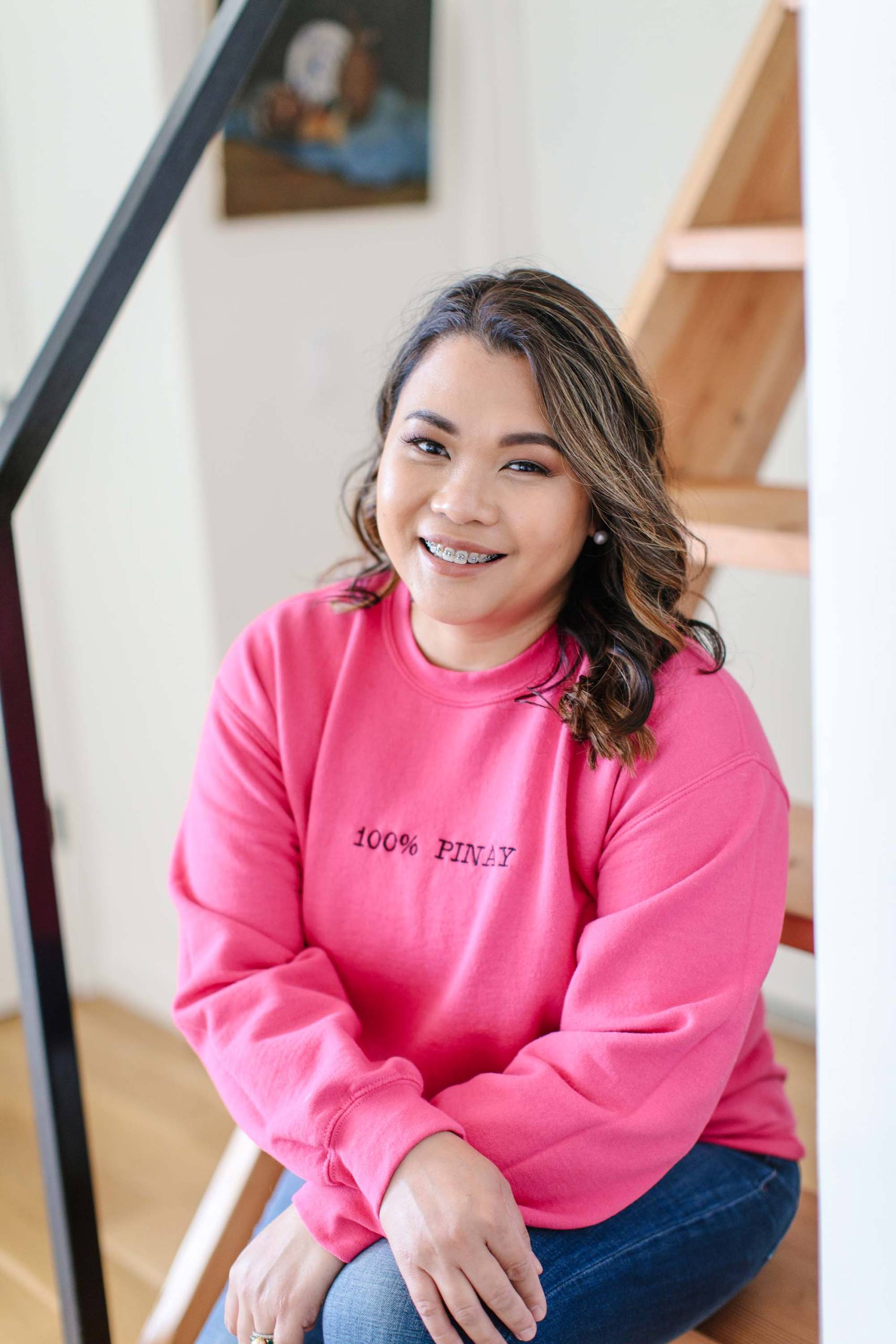 A Filipina woman seated on a staircase poses for a portrait. The text on the front of her pink sweatshirt reads, "100% Pinay".