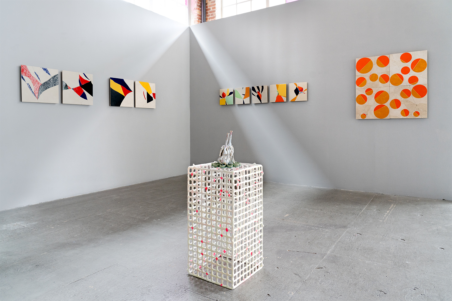 Fractured and multicolored tile pieces on light gray walls, in foreground a sculpture made of white gridded material with a ceramic piece on top