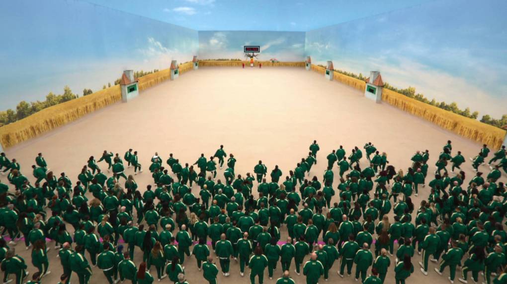 Hundreds of people wearing matching green tracksuits stand at one end of a vast room with sandy colored floor and walls painted to resemble the sky.