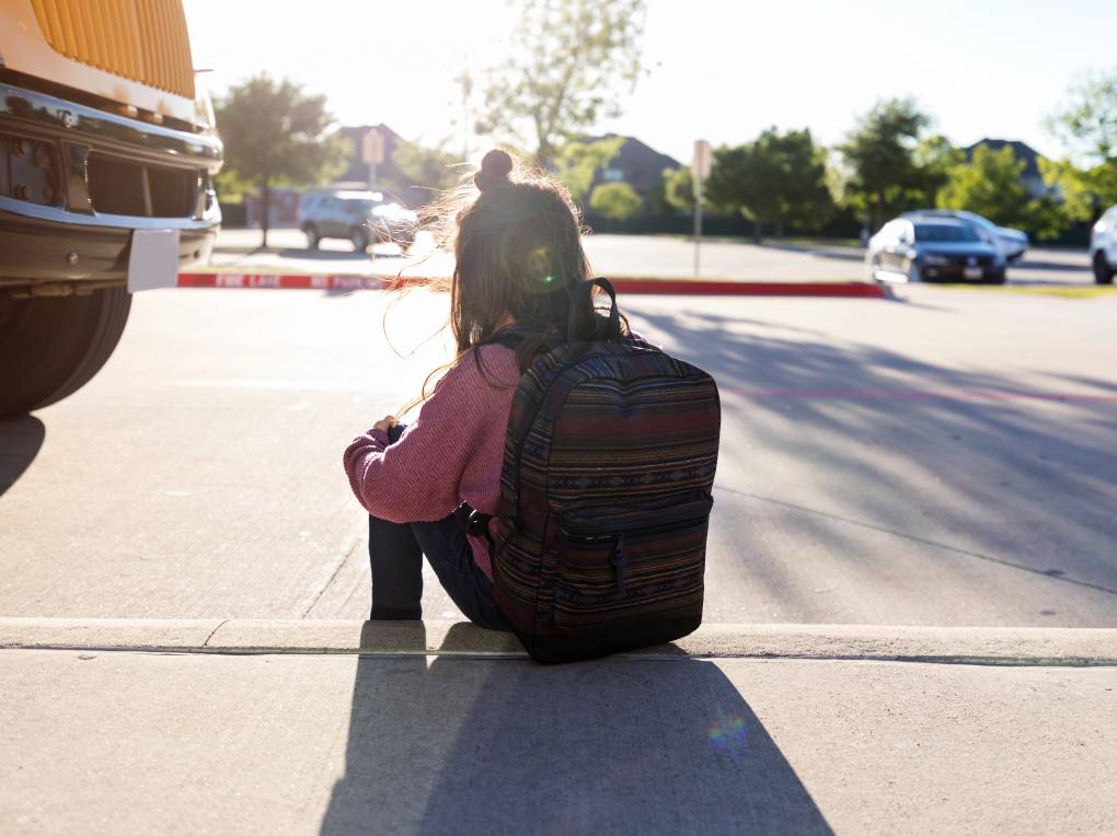 An unknown schoolgirl with a backpack hugs her knees and sits on the curb by herself at a bus stop, while the bus is parked nearby.