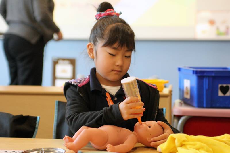 A child with hair in a bun feeds a doll in a classroom.