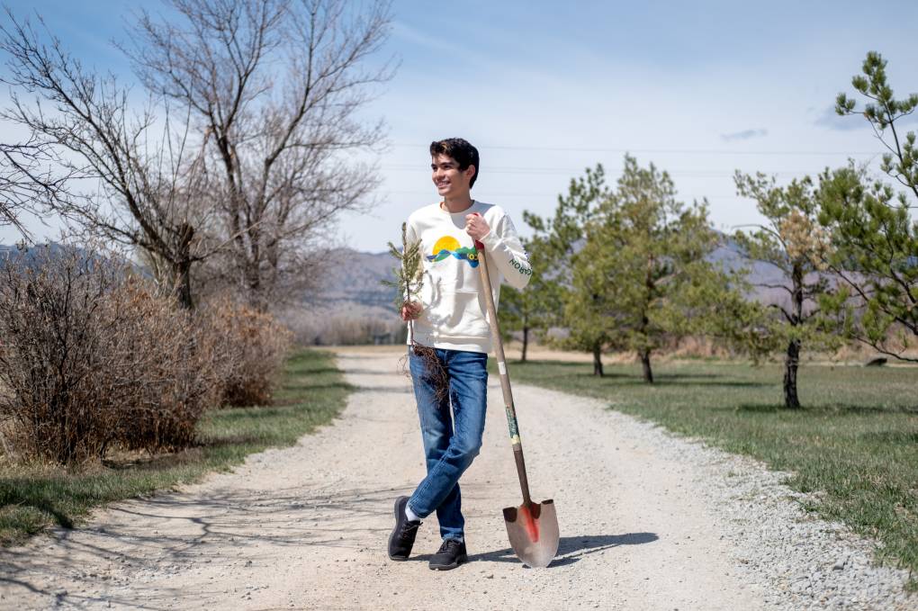 Teenage boy in jeans, sneakers and sweathshirt stands on a dirt path with a shovel and baby tree in hand. Trees line either side of the path and mountains are in the distance..