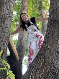 A teenage girl in white overalls with pink designs and a black short-sleeved shirt stands in a tree with arms streteched between two tree trunks.