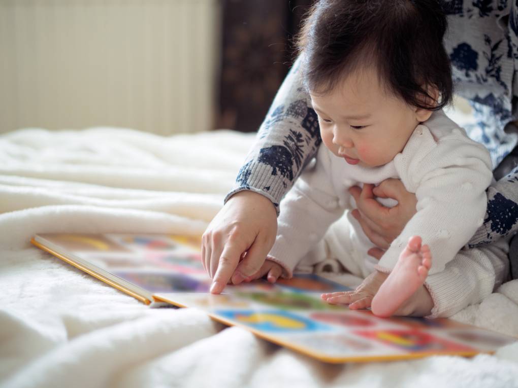 baby girl reading book with caregiver arms holding her and one hand pointing to the book