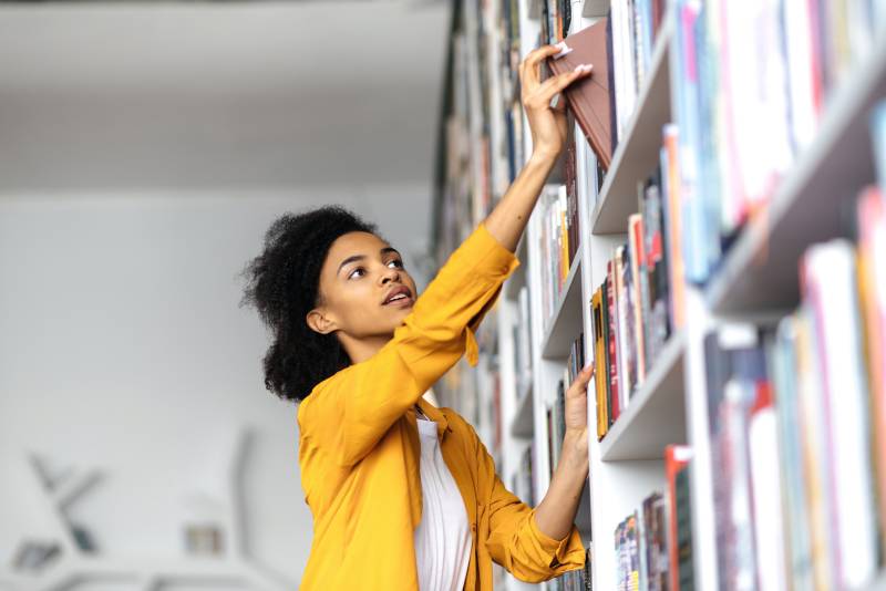 African american female student stands in university library, looking for a book. A pretty clever girl searching information for a project, choosing book at library bookshelf, education concept