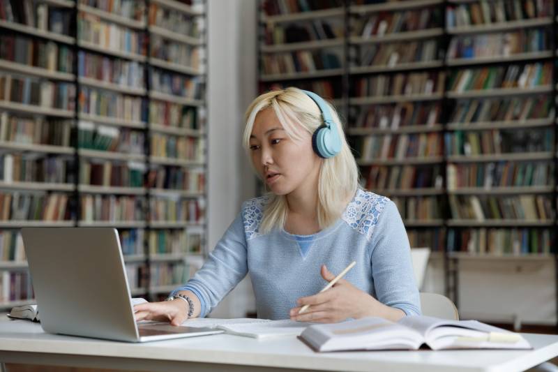 Asian female college student with blonde hair wearing headphones and using a laptop at the library