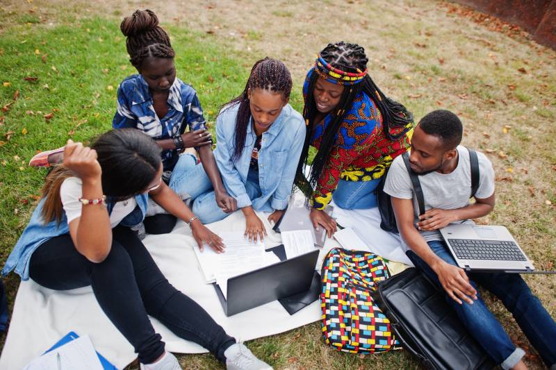 group of 5 Black student with notebooks and laptops study and chat outside on grass
