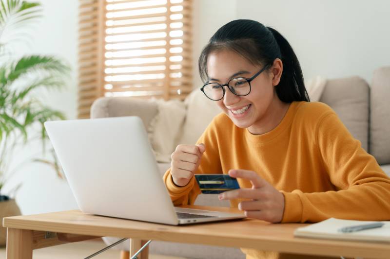 young woman buying online with a credit card while sitting in the living room morning. Women is using a computer laptop and doing online transactions at home
