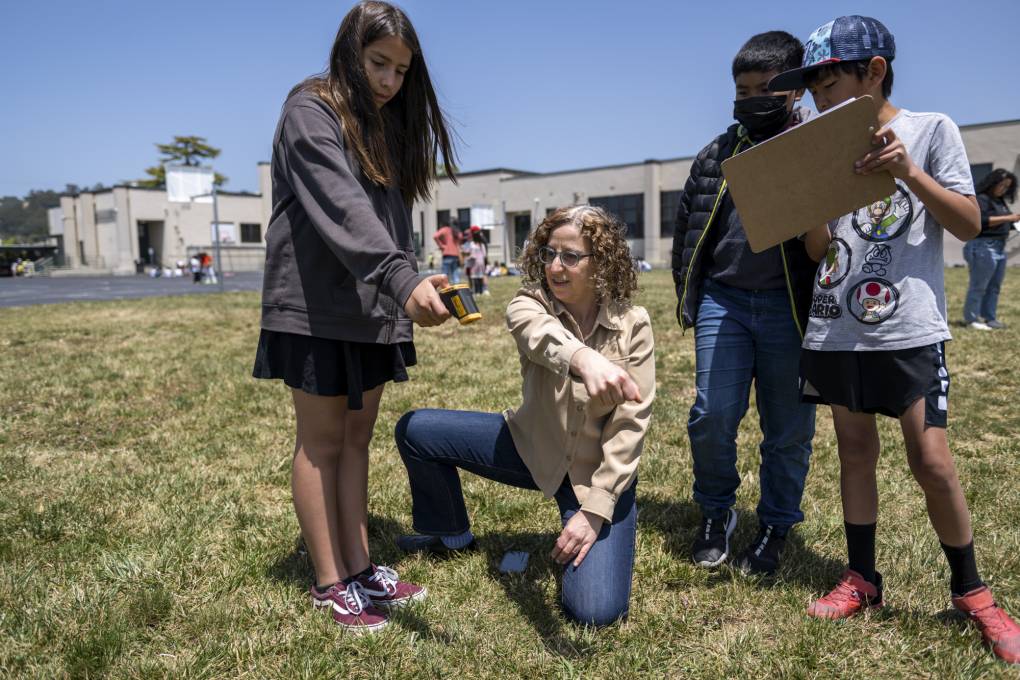 In a schoolyard, one child points a thermometer at the grass, while another writes on a clipboard, and a third watches. An adult woman kneels and points.