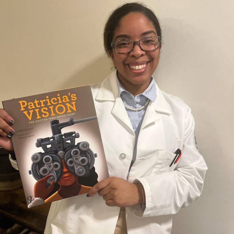 Lafayette holds up Patricia's Vision: The Doctor Who Saved Sight while she is dressed as Dr. Patricia Bath, a groundbreaking ophthalmologist who pioneered laser surgery.