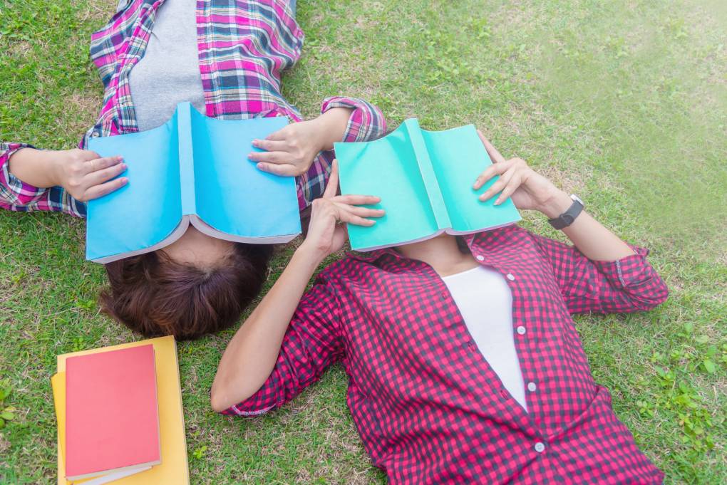 Two students with stack of books close their face, laying in the grass.