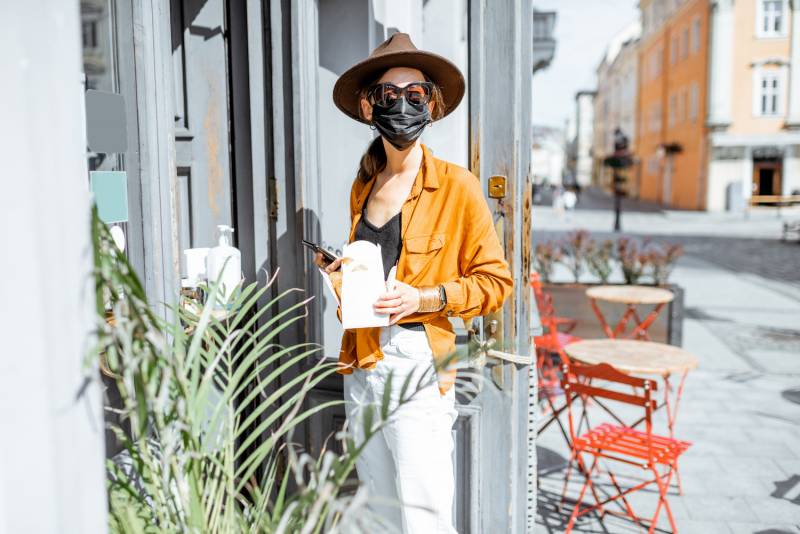 Portrait of a young woman in facial protective mask standing with a takeaway food near the cafe entrance outdoor. Concept of a new social rules for business after coronavirus pandemic
