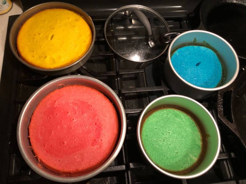 Cake tins on a stove with different colors. One is yellow, one is res, one is green and one is blue. There is also a pot lid on the stove