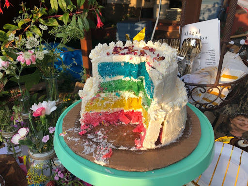 A rainbow cake that has been cut into with white buttercream frosting on the outside. It's on a teal cakestand with a table surrounded by flowers, gold-stripped plates, cutlery and wedding programs.