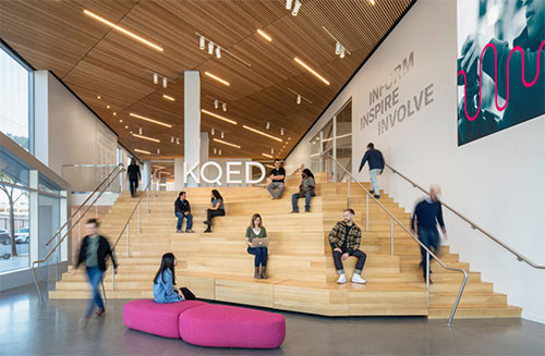 People sit and walk on the wide wooden stairs and built-in seats in the KQED lobby