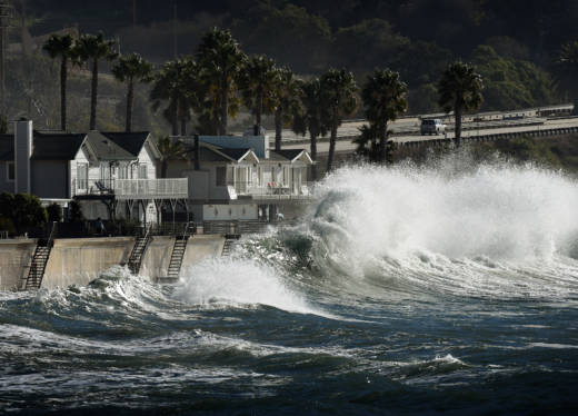 A large wave crashes onto seaside houses.
