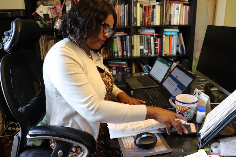 A woman with coffee skin and black hair, wearing a white sweater, sits at a desk in front of two laptops. This is Teron McGrew at her home office in North Oakland. Rows of books, with books stacked on top of them, fill her bookshelf.