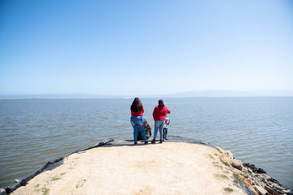 Two women with black hair stand at the end of light sand colored patch of ground looking out onto open water, with blue sky above.