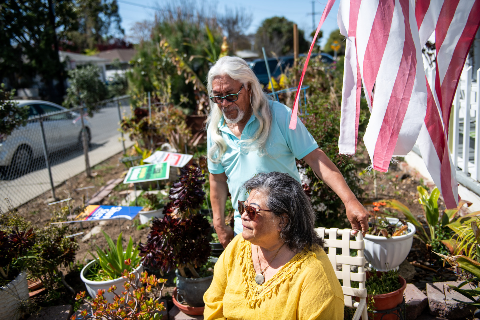 A woman in a marigold colored sundress and a long white-haired man in light blue stand and sit in their front yard surrounded by plants and an American Flag waves in the wind. 