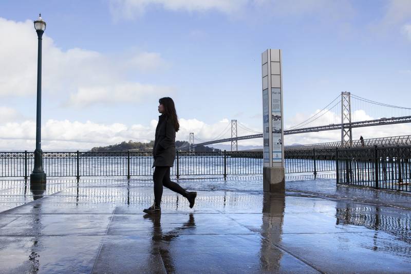 a person walks along wet concrete, with the bay bridge behind and cloudy blue skies above.