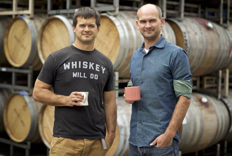 Ryan Harms and Eric Harms stand in front of big oak wine barrels. 