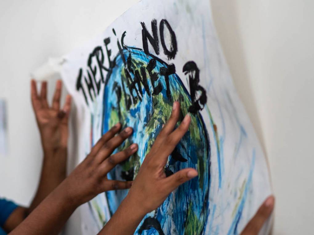 The hands of several children hold up a handmade sign with a painting of Earth and copy that reads "There's No Planet B."