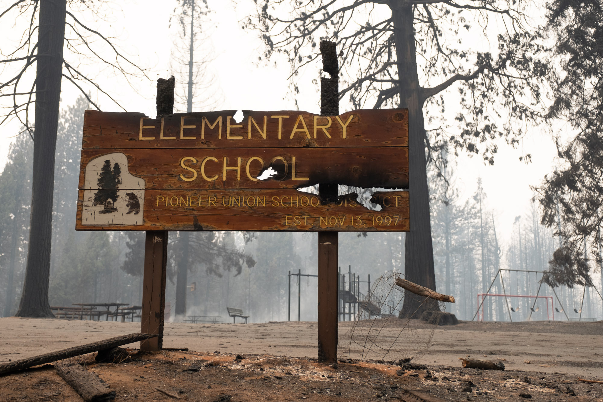 A burned entrance sign at Walter Tyler Elementary school with charred playground equipment in the background. 