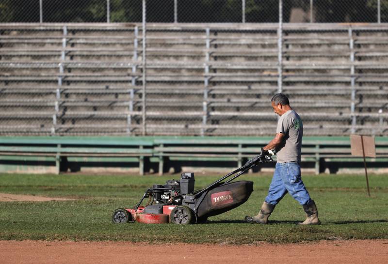 A park worker pushes a large, red and black lawn mower in front of bleachers on baseball field.