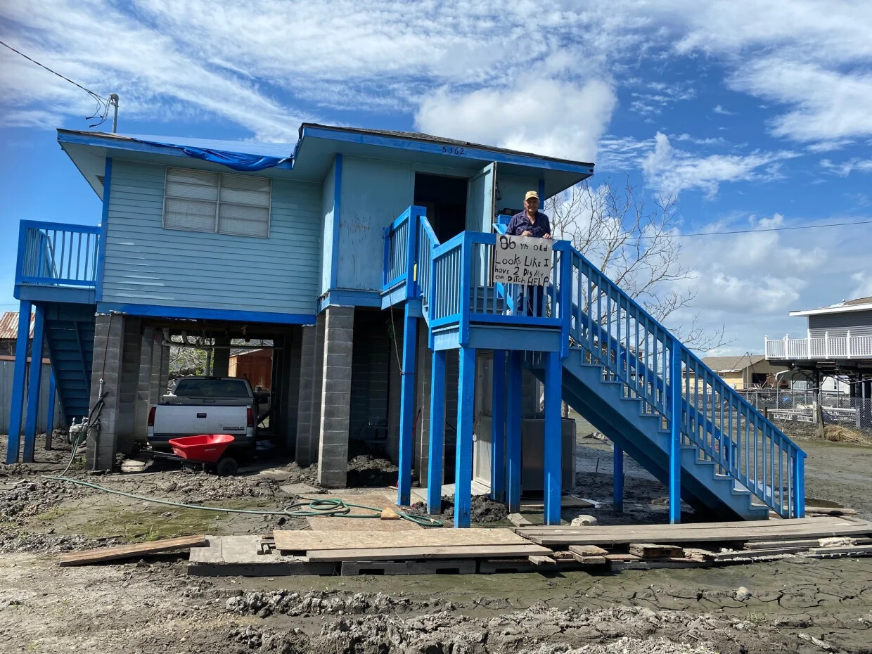 A man stands on the steps outside of a bright blue house, on the second floor landing.