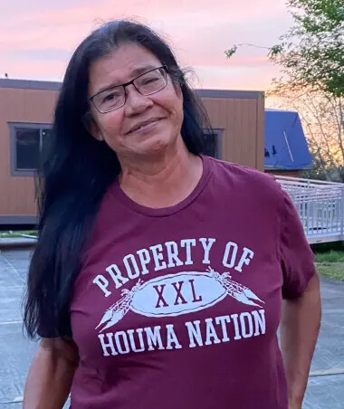 A woman with black and gray hair, square dark-rimmed glasses, and a merlot-colored shirt that says "property of Houma Nation" in capital letters. 